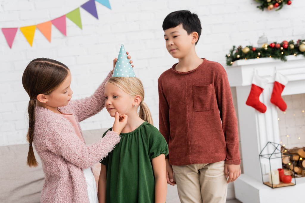 Dívka nošení mejdan čepice na přítele v blízkosti asijské chlapec během narozeninové párty doma  - Fotografie, Obrázek