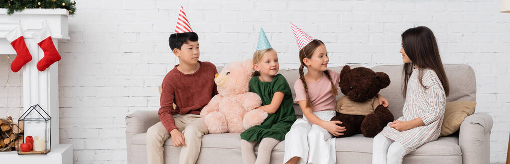 Мультиэтнические дети в шапочках для вечеринок празднуют день рождения возле мягких игрушек и рождественского декора, баннер  - Фото, изображение