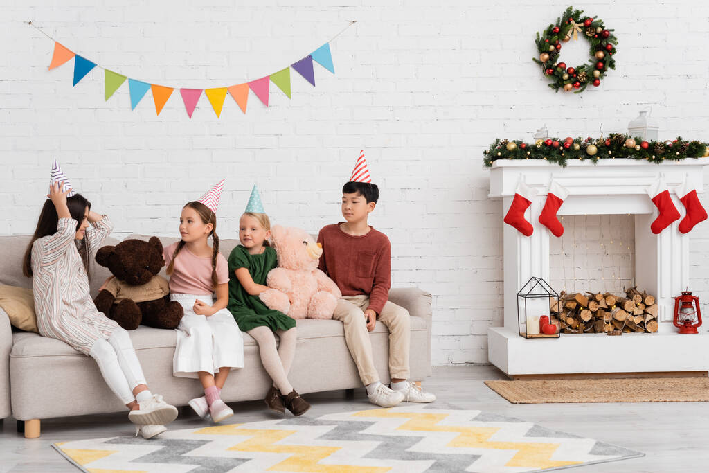 Vidám interracial gyerekek party sapkában ül mellett plüssmacik és karácsonyi dekoráció otthon  - Fotó, kép