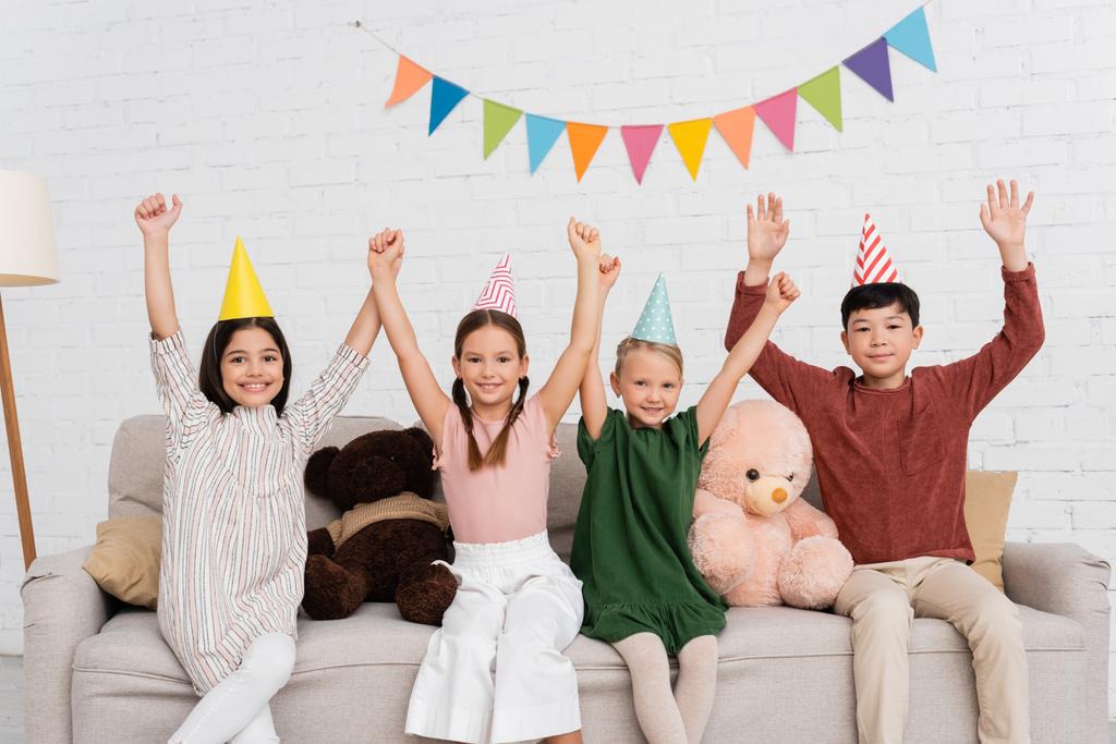 Excited multiethnic kids in party caps looking at camera on couch during birthday party at home  - Photo, Image