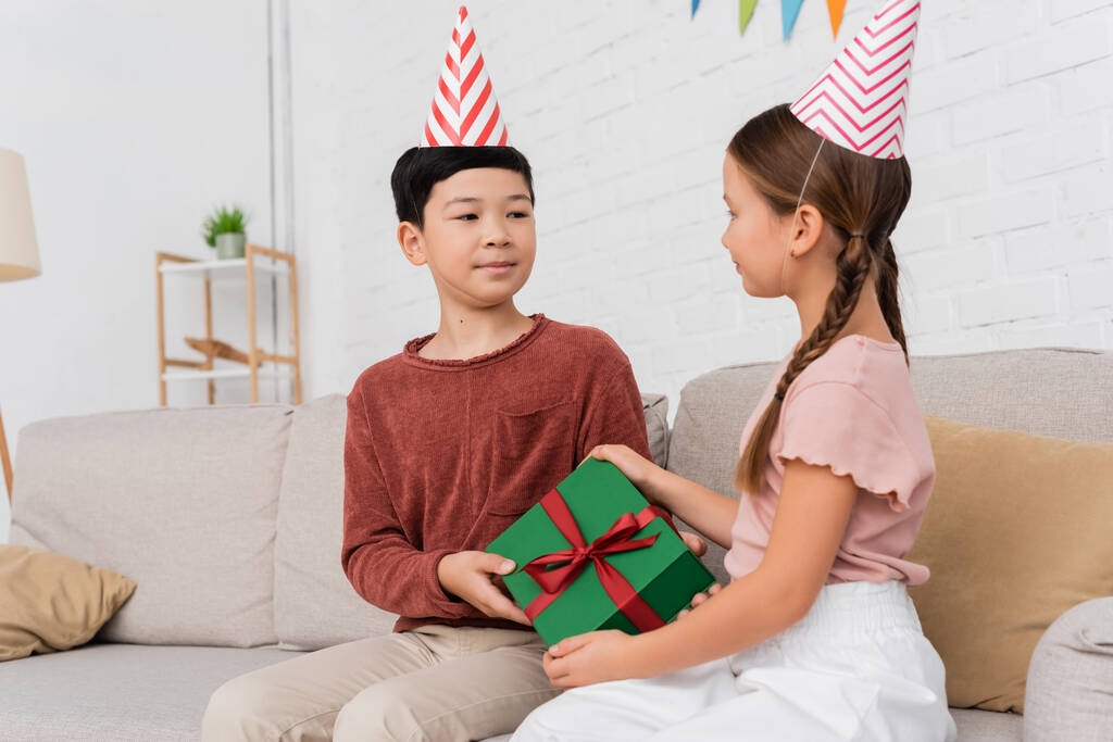 Parti şapkalı Asyalı çocuk evde doğum günü partisinde arkadaşına hediye veriyor.  - Fotoğraf, Görsel