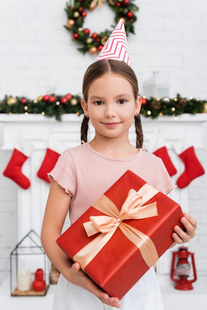Retrato de niño en gorra de fiesta sosteniendo caja de regalo cerca de la decoración borrosa de Navidad en casa  - Foto, Imagen