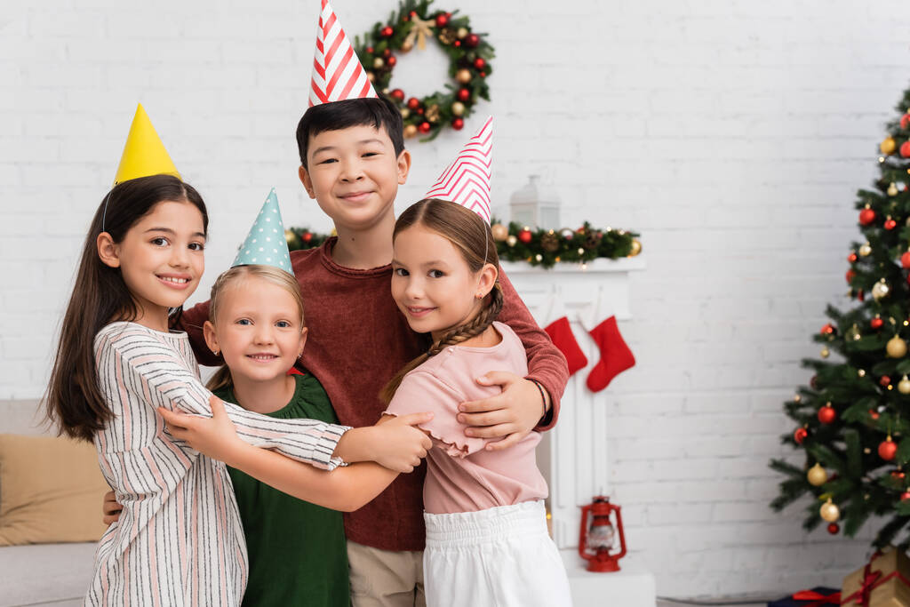 Souriant enfants multiethniques dans des casquettes de fête étreignant pendant l'anniversaire et la célébration du Nouvel An à la maison  - Photo, image