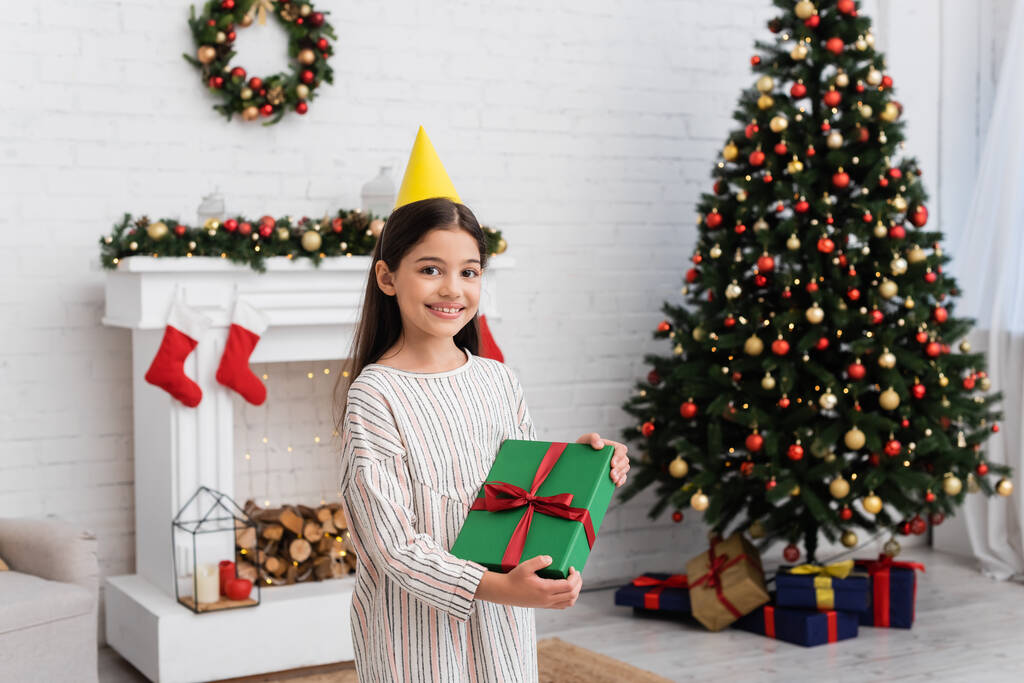 Happy girl in party cap holding gift box közelében elmosódott karácsonyfa és kandalló otthon  - Fotó, kép