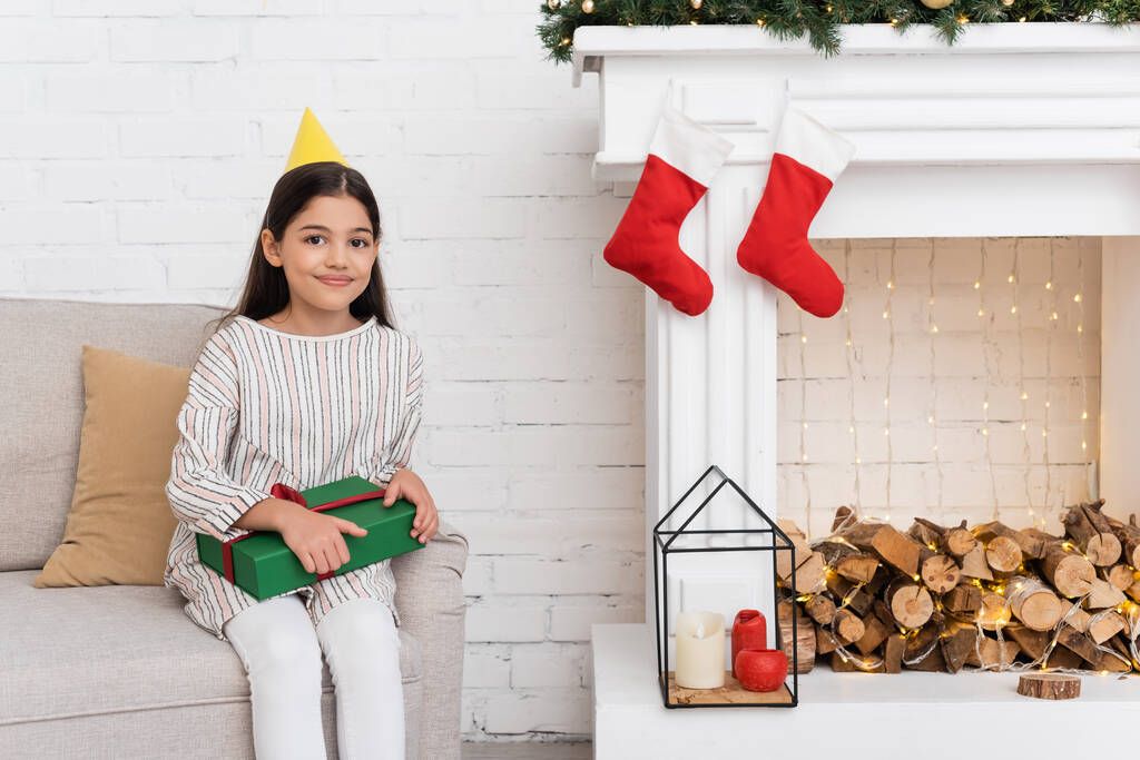 Uśmiechnięta dziewczyna w czepku party trzyma obecny na kanapie w pobliżu kominka z świątecznym wystrojem w domu  - Zdjęcie, obraz
