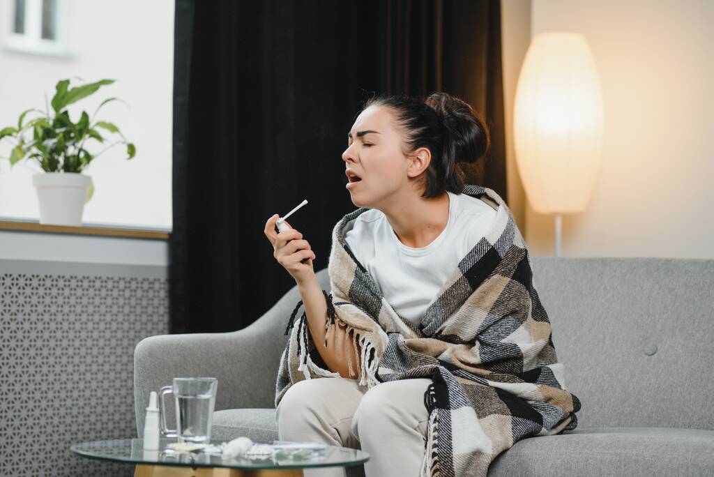 sairas nuori nainen lämpimissä vaatteissa ruiskuttamalla kurkkua istuessaan sohvalla kotona
 - Valokuva, kuva