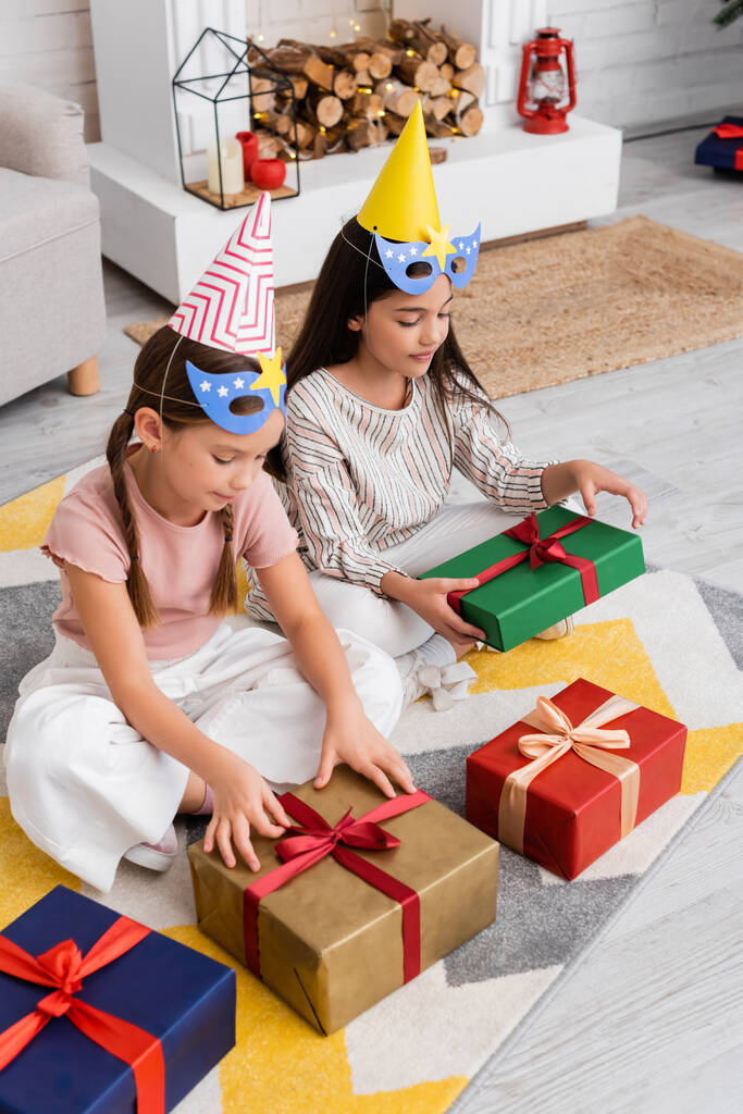 Вид под высоким углом на девочек в кепках и масках, смотрящих на подарки во время дня рождения дома  - Фото, изображение