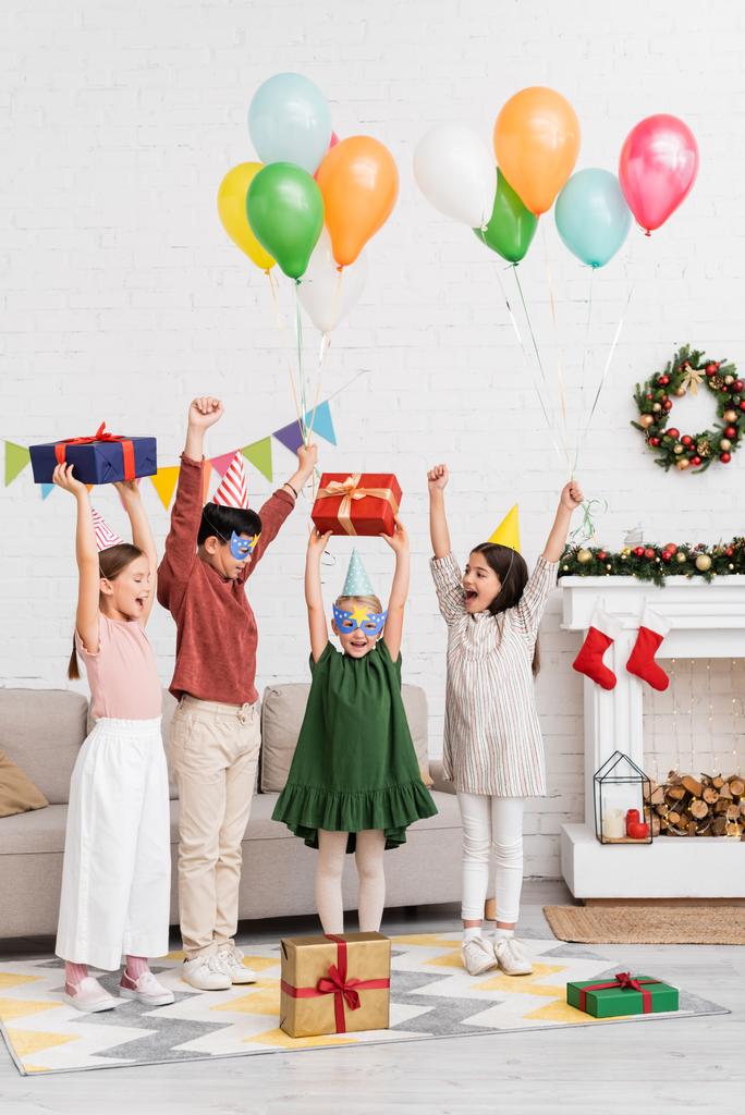 Bambini multietnici eccitati in cappelli e maschere che tengono palloncini vicino all'amico con regalo di compleanno e decorazioni natalizie a casa  - Foto, immagini