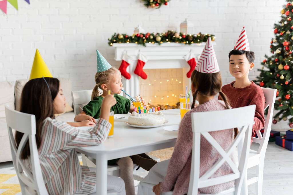 Niños interraciales en gorras de fiesta sentados cerca de pastel de cumpleaños y jugo de naranja en casa en invierno  - Foto, imagen