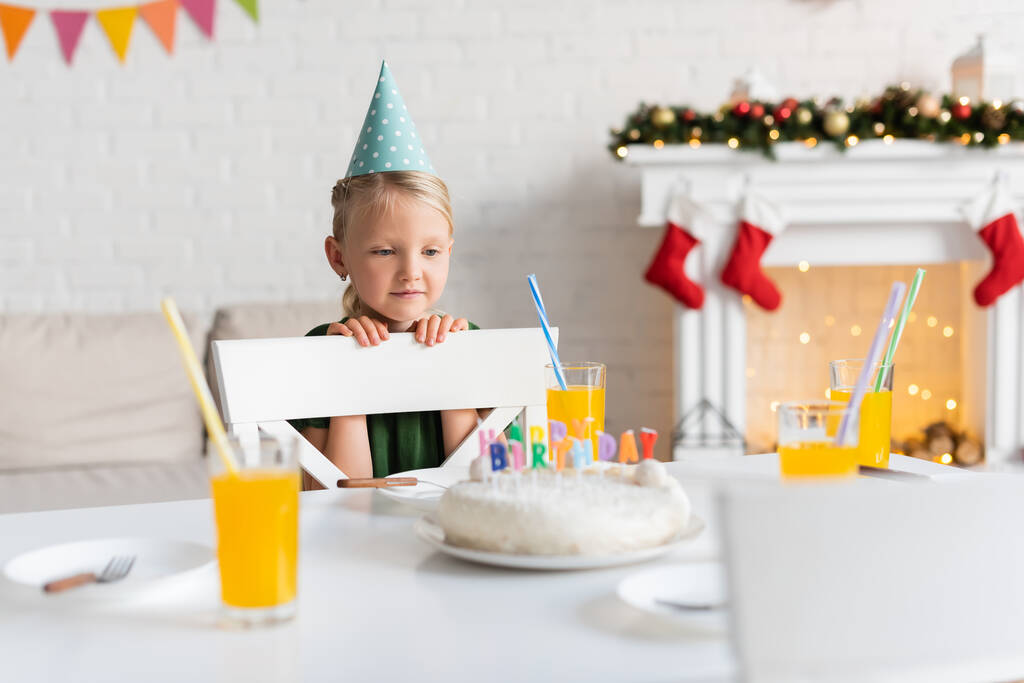 Kind mit Partymütze schaut zu Hause auf Gläser mit Orangensaft und Geburtstagstorte  - Foto, Bild