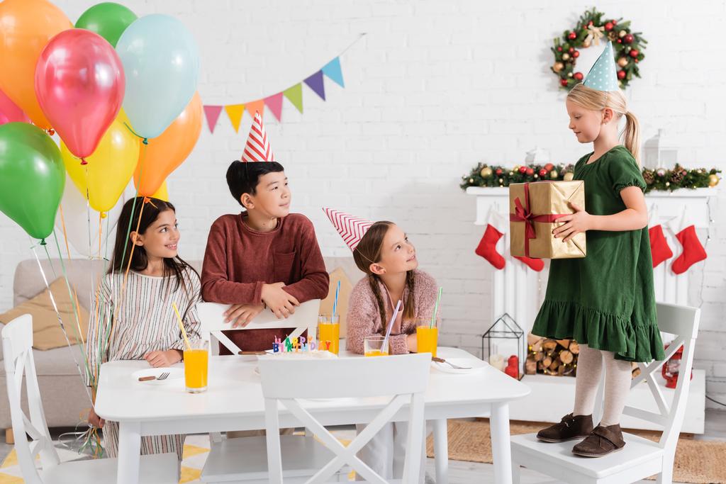 Межрасовые дети в шапочках для вечеринок держат воздушные шары рядом с другом с подарком и праздничным тортом дома  - Фото, изображение