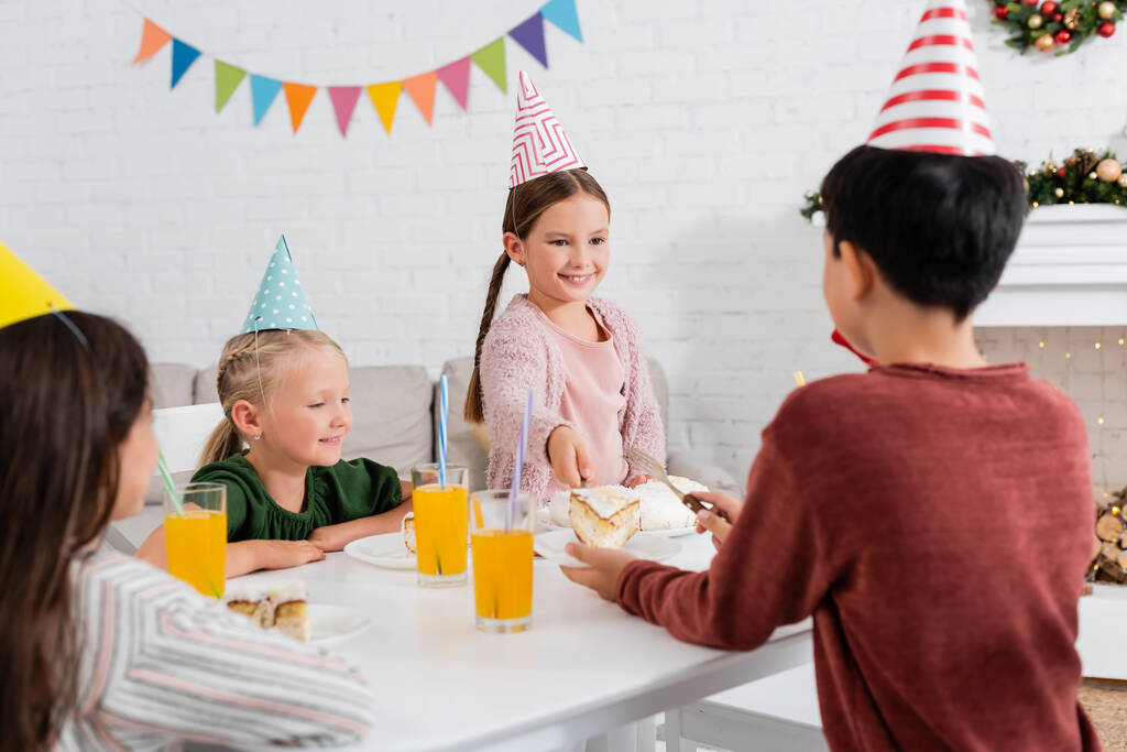 Kid in party cap giving birthday cake to blurred friend near children at home  - Fotografie, Obrázek