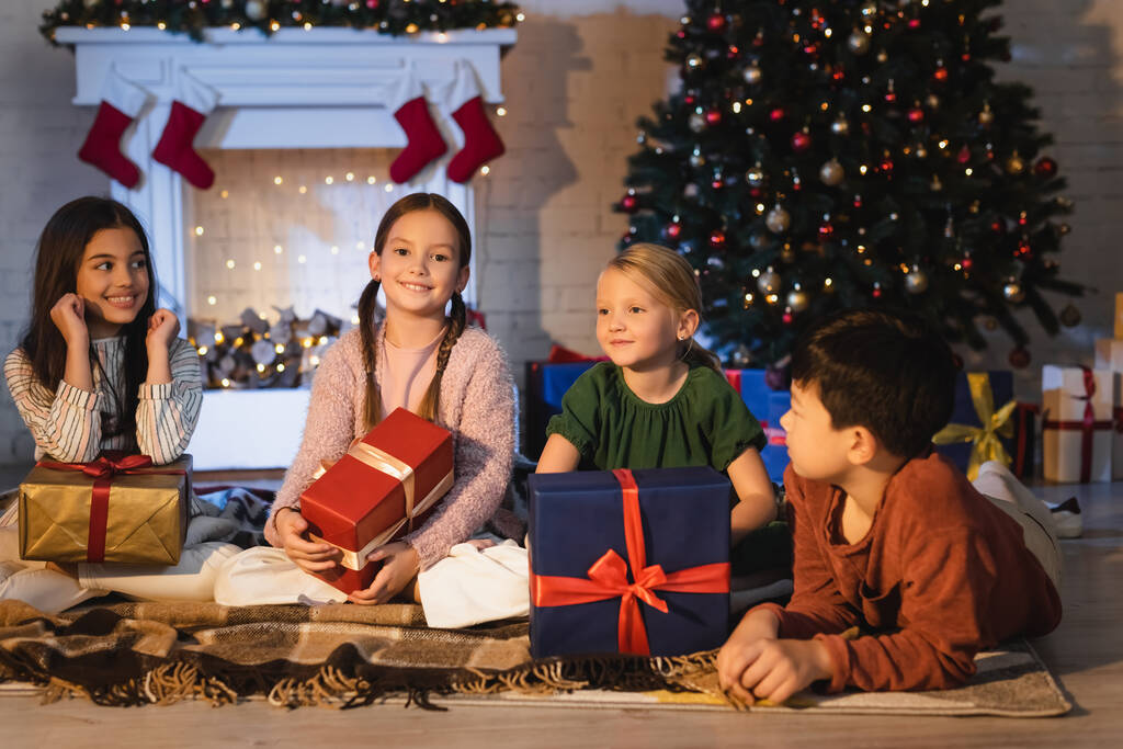 Niños multiétnicos felices sosteniendo regalos en la manta cerca de la chimenea y el árbol de Navidad en casa  - Foto, imagen