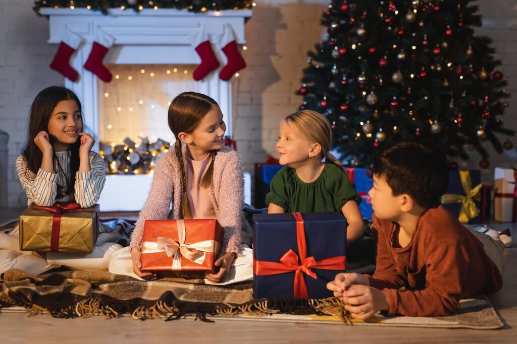Усміхнені багатоетнічні діти розмовляють біля різдвяних подарунків і каміна вдома
  - Фото, зображення