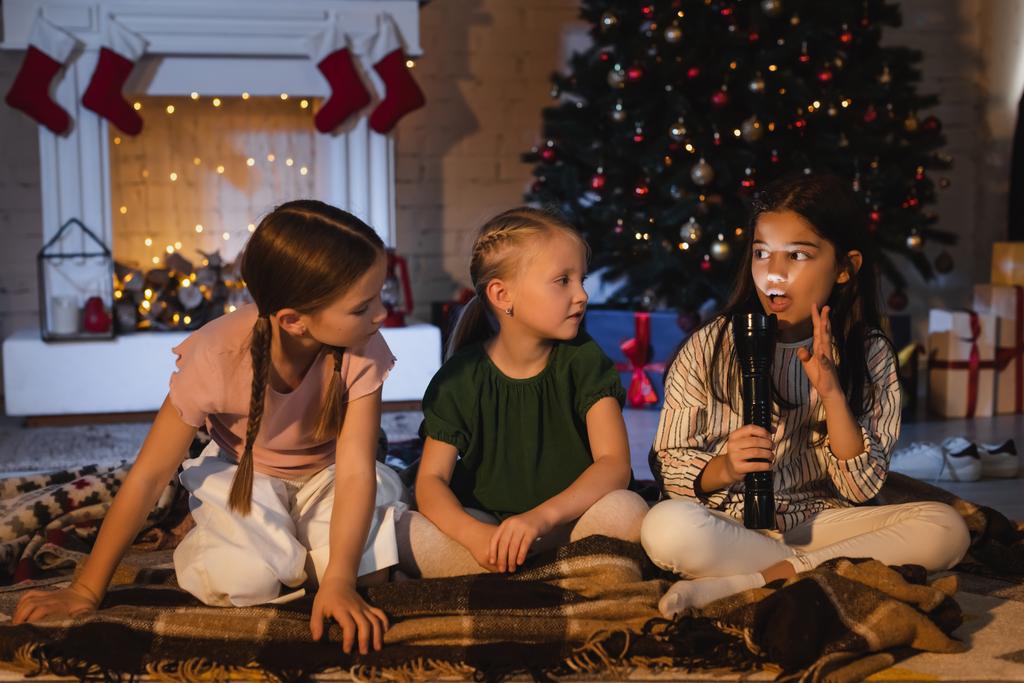 Κορίτσι με φακό μιλάει κοντά σε φίλους σε κουβέρτα και θολή χριστουγεννιάτικο δέντρο στο σπίτι  - Φωτογραφία, εικόνα