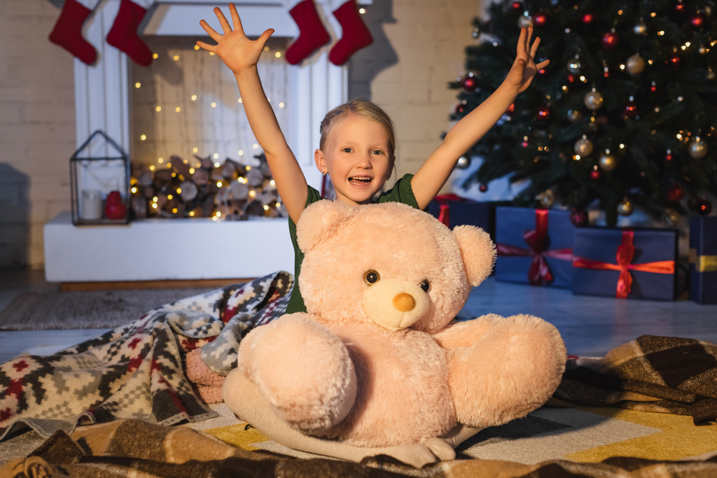 Ragazza positiva guardando la fotocamera vicino orsacchiotto sulla coperta e sfocato albero di Natale a casa  - Foto, immagini