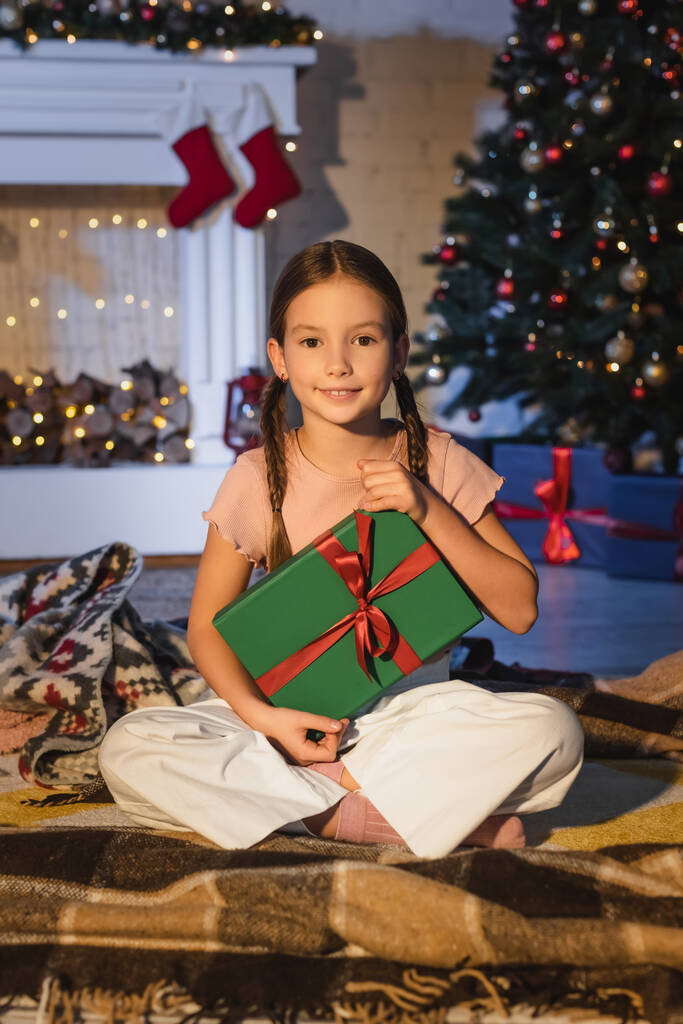 クリスマスツリーの近くの毛布と自宅で暖炉に座っている間にプレゼントを保持している未成年の子供  - 写真・画像