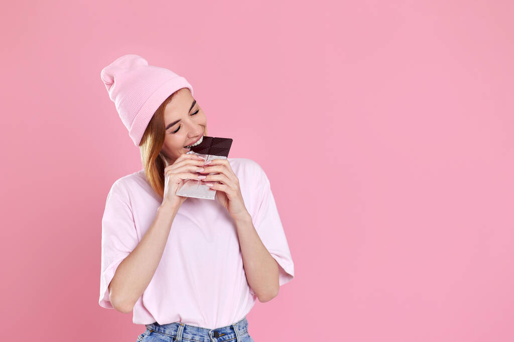 Hermosa mujer joven en camiseta casual mordiendo deliciosa barra de chocolate aislado sobre fondo rosa pastel - Foto, Imagen
