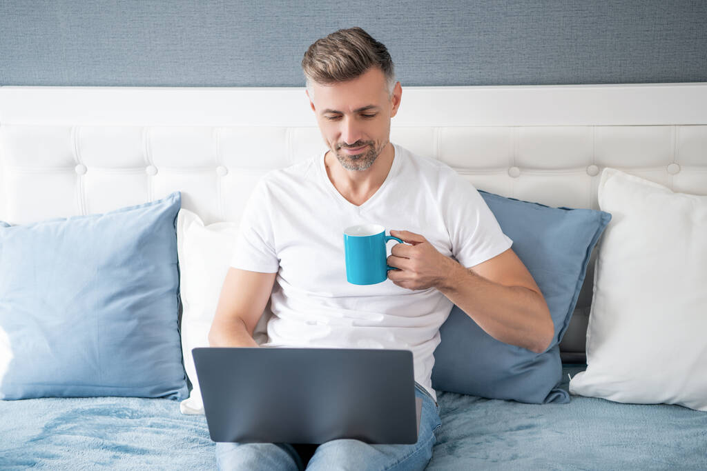 ώριμος άντρας που χρησιμοποιεί φορητό υπολογιστή στο κρεβάτι με καφέ. - Φωτογραφία, εικόνα