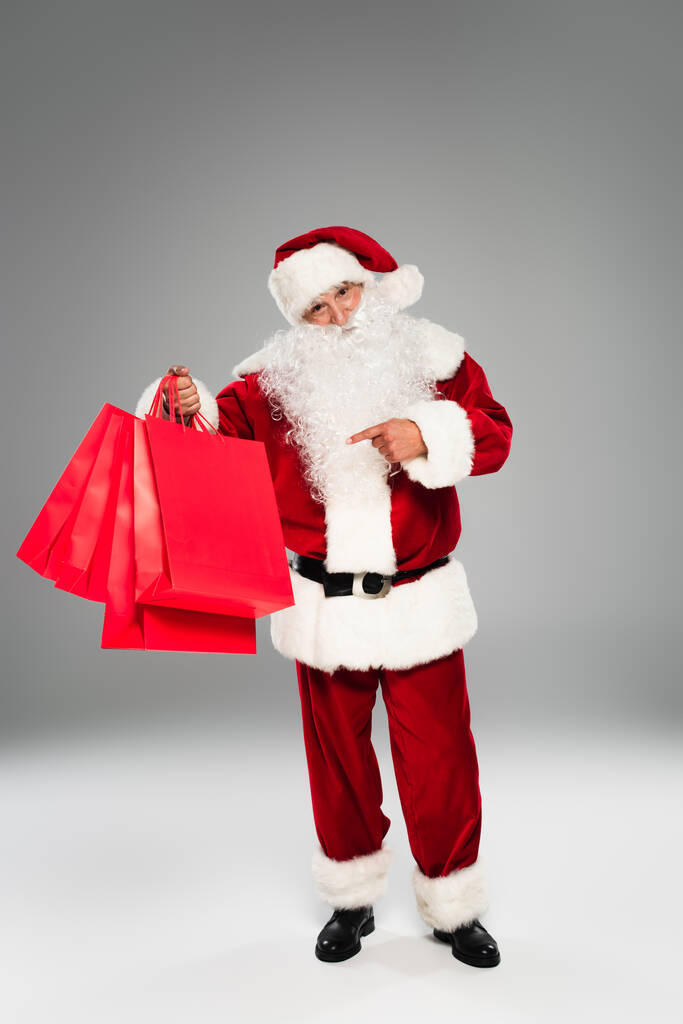 Santa Claus ukazuje na červené nákupní tašky na šedém pozadí  - Fotografie, Obrázek
