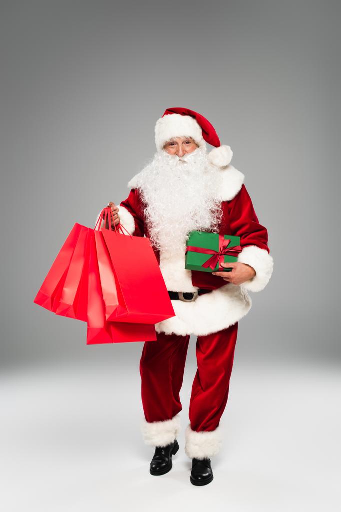 Повна довжина Санта-Клауса тримає червоні сумки для покупок та подарунок на сірому фоні
  - Фото, зображення