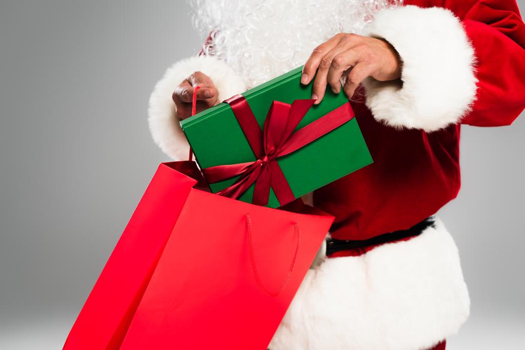 Vista cortada de Papai Noel barbudo colocando presente em saco de compras isolado em cinza  - Foto, Imagem