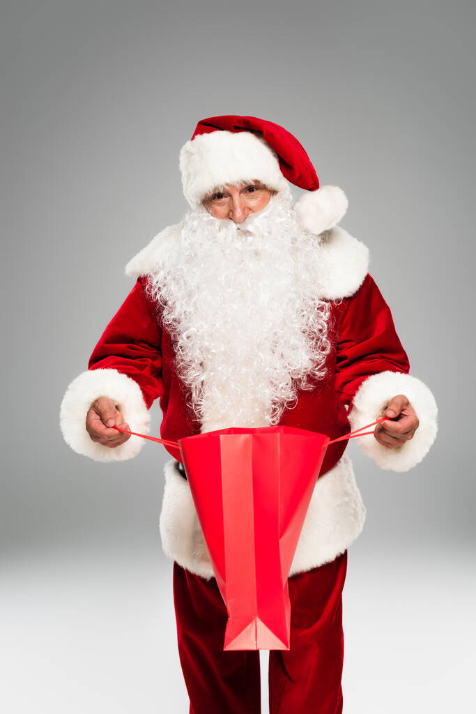 Santa Claus en sombrero abriendo bolsa de compras y mirando a la cámara aislada en gris  - Foto, imagen