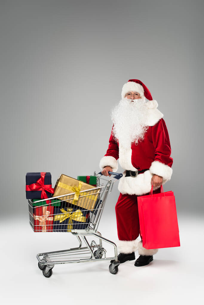 Ojciec Boże Narodzenie trzymając torby na zakupy w pobliżu koszyka z prezentami na szarym tle  - Zdjęcie, obraz