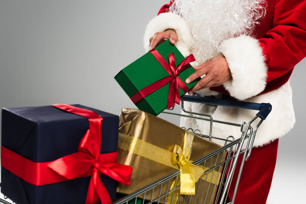 Gesneden weergave van de kerstman in kostuum zetten geschenkdoos in winkelwagen geïsoleerd op grijs  - Foto, afbeelding