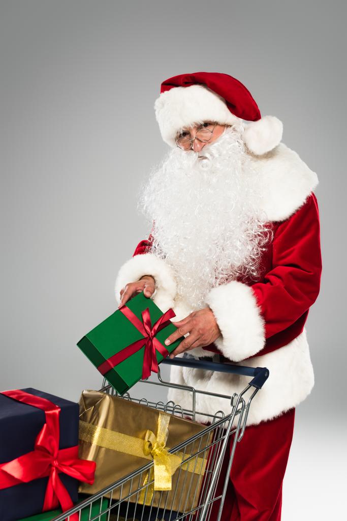 Père Noël en costume tenant boîte cadeau près de présent dans le panier isolé sur gris  - Photo, image