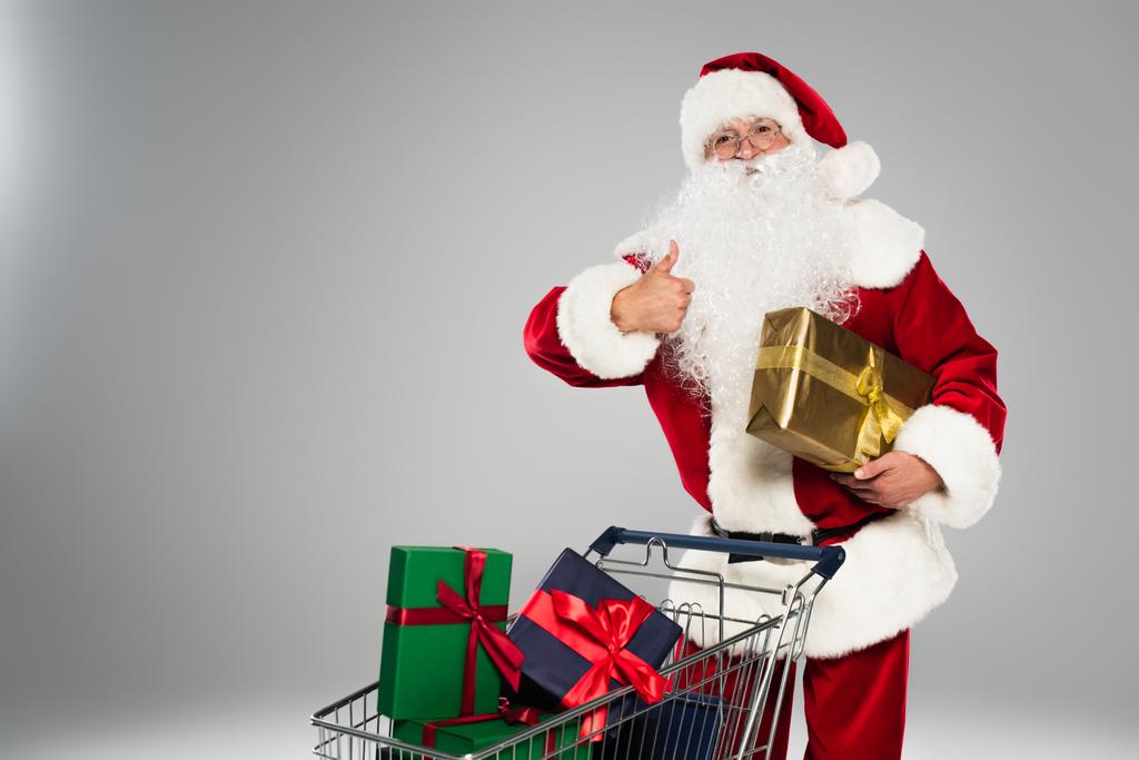 Święty Mikołaj pokazując jak gest i trzymając obecny w pobliżu koszyka handlowego odizolowany na szaro  - Zdjęcie, obraz
