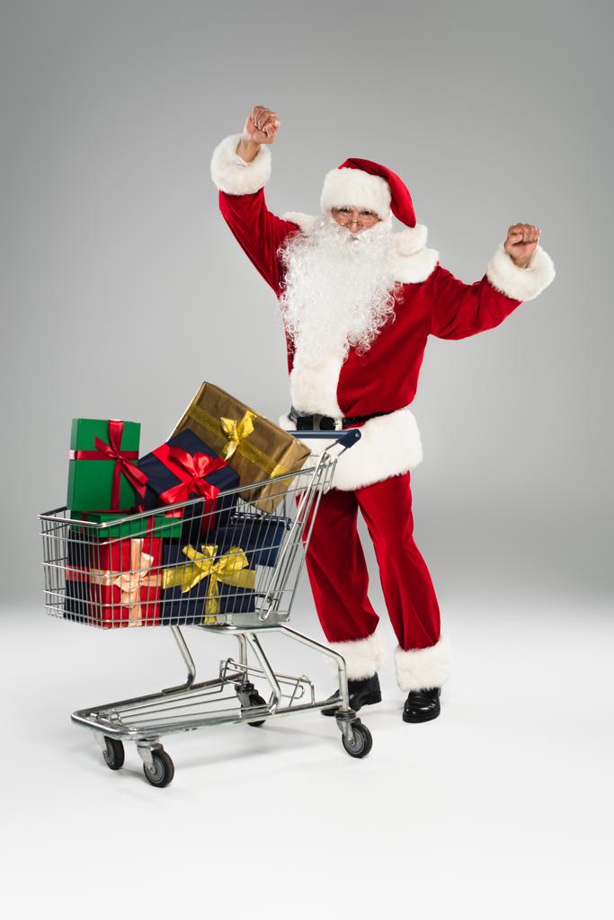 Pełna długość podekscytowany Święty Mikołaj stojący w pobliżu koszyka z prezentami na szarym tle  - Zdjęcie, obraz