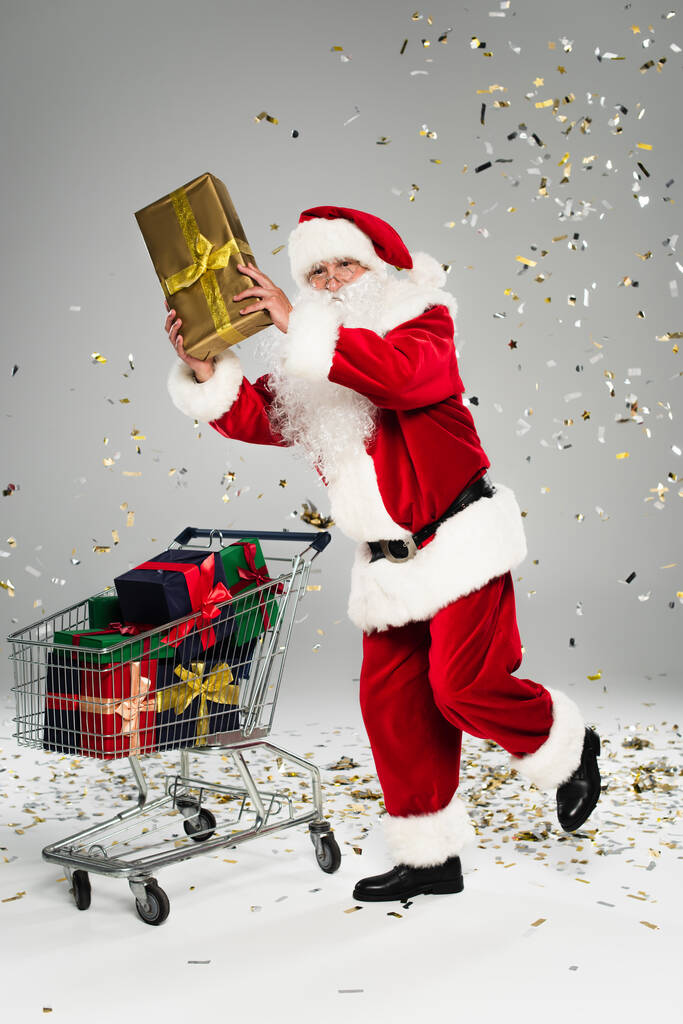 Kerstman holding aanwezig in de buurt van winkelwagen en confetti op grijze achtergrond  - Foto, afbeelding