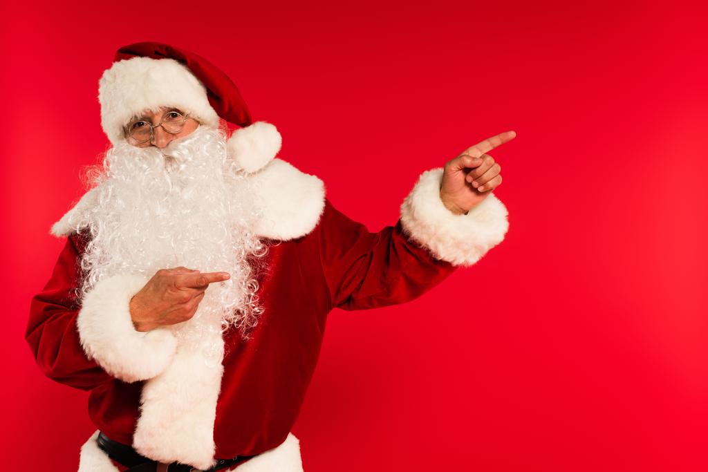 Санта-Клаус в очках указывает пальцами и смотрит на камеру, изолированную на красном  - Фото, изображение