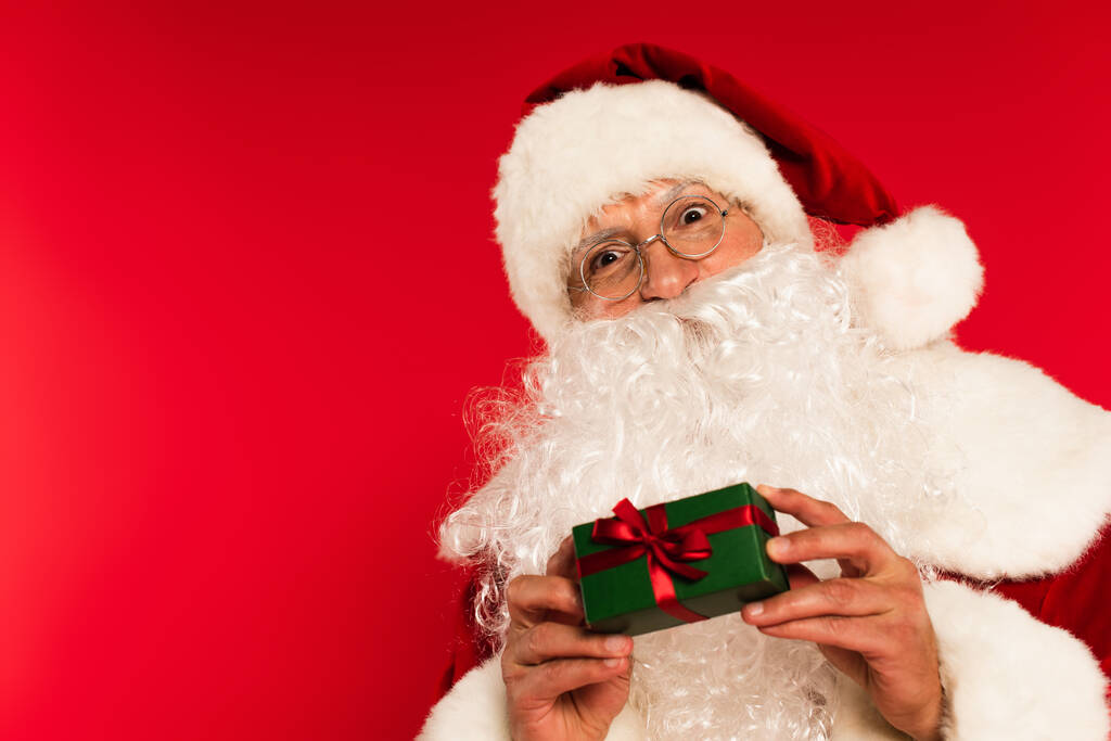 Γενειοφόρος πατέρας Χριστούγεννα εκμετάλλευση μικρό κουτί δώρου απομονώνονται σε κόκκινο  - Φωτογραφία, εικόνα