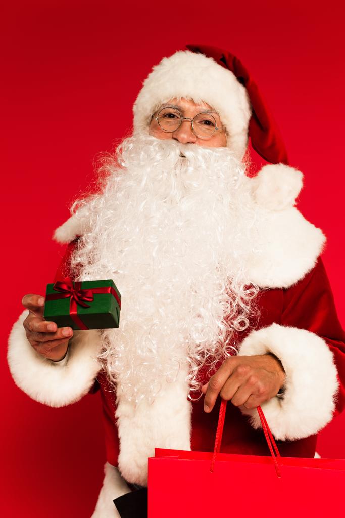 Santa Claus κρατώντας τσάντα αγορών και μικρό δώρο σε κόκκινο φόντο  - Φωτογραφία, εικόνα
