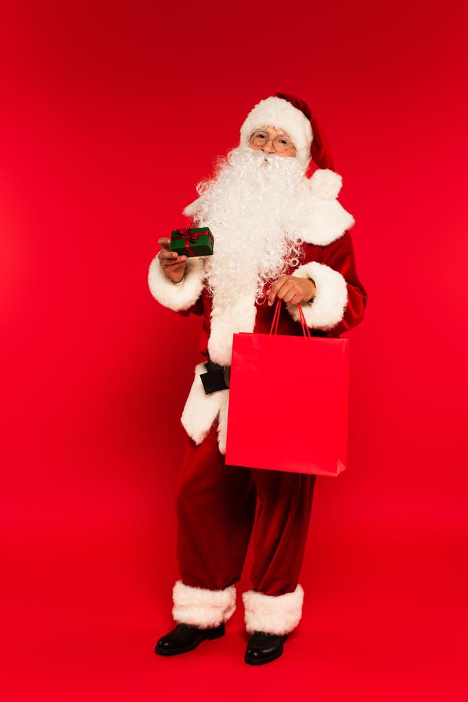 Pleine longueur de santa claus tenant sac à provisions et petite boîte cadeau avec arc sur fond rouge  - Photo, image