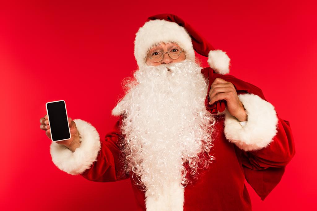 Отец Рождество с мешком держа смартфон с пустым экраном на красном фоне  - Фото, изображение