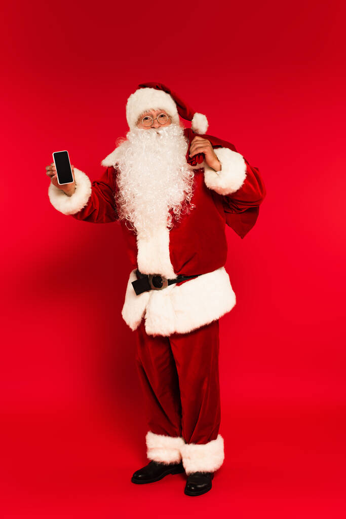Kerstman in kostuum houden zak en mobiele telefoon met leeg scherm op rode achtergrond  - Foto, afbeelding
