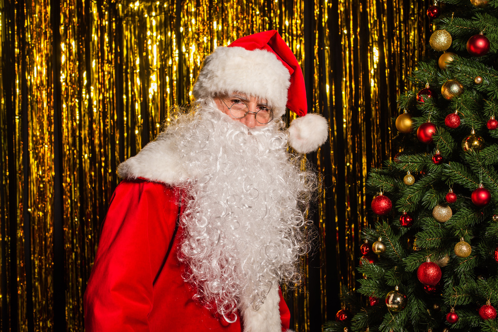 Weihnachtsmann in Kostüm und Brille blickt in die Kamera neben Weihnachtsbaum und goldenem Lametta  - Foto, Bild