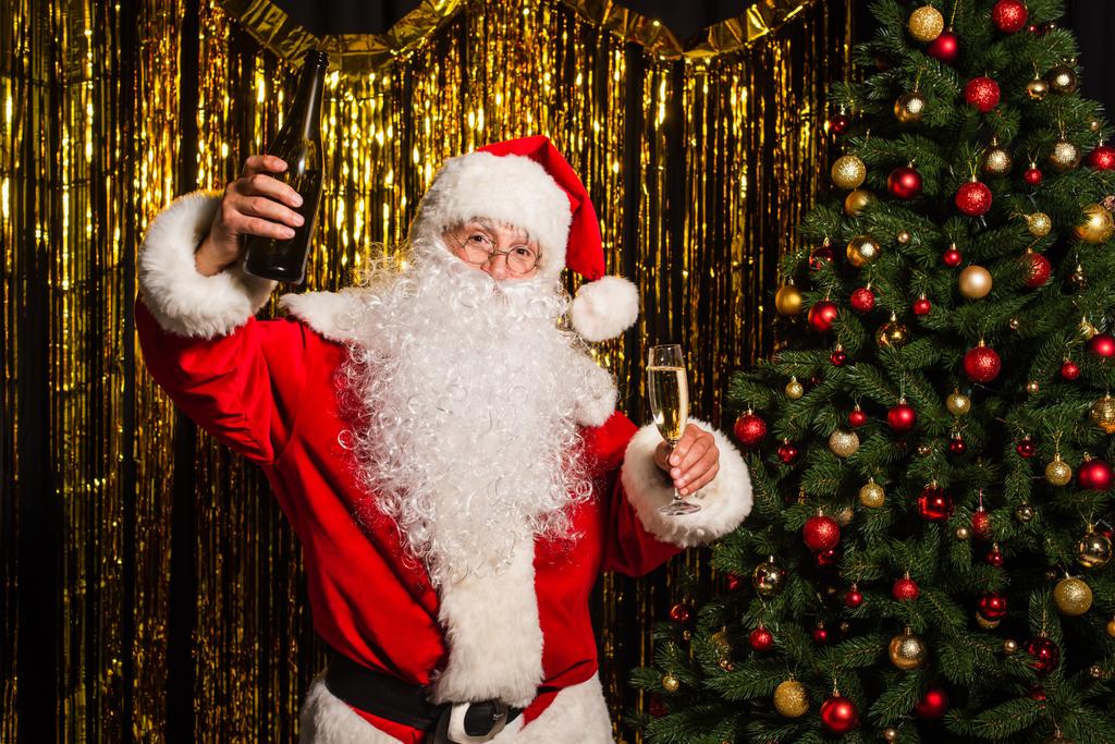 Weihnachtsmann hält Flasche und Glas Champagner in der Nähe von Kiefer und Lametta  - Foto, Bild