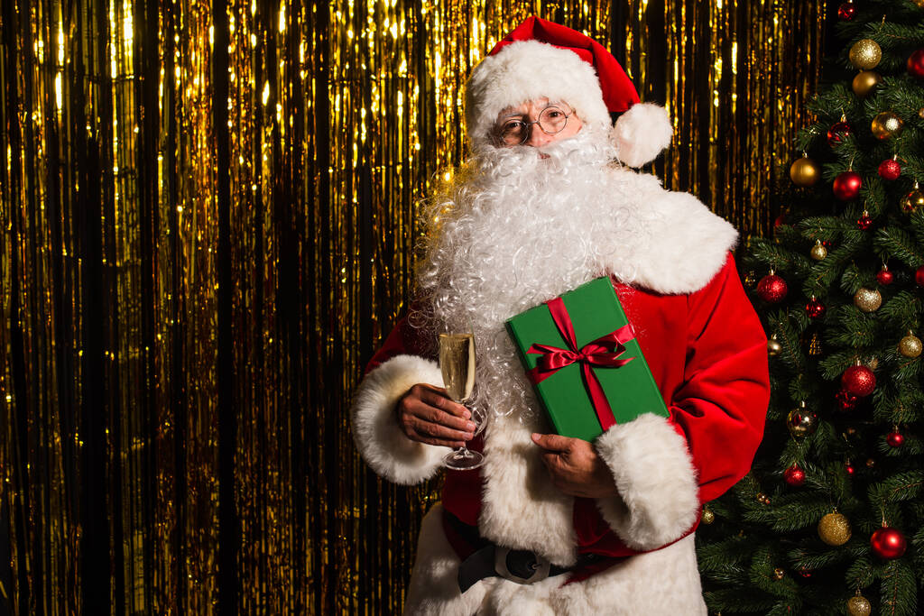 Weihnachtsmann hält Geschenk und Champagner neben Weihnachtsbaum und Lametta  - Foto, Bild