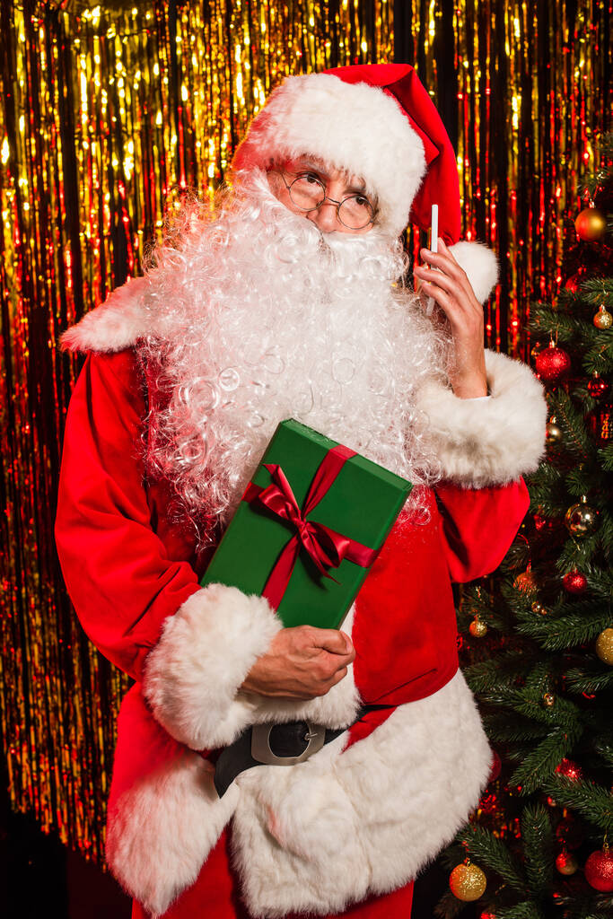 Santa claus holding smartphone and gift near decorated christmas tree and tinsel  - Photo, Image