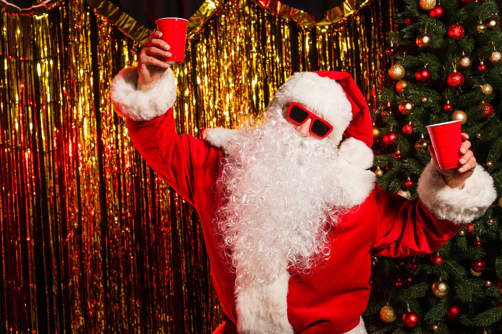 Santa claus in sunglasses holding disposable cups near christmas tree and tinsel  - Photo, Image