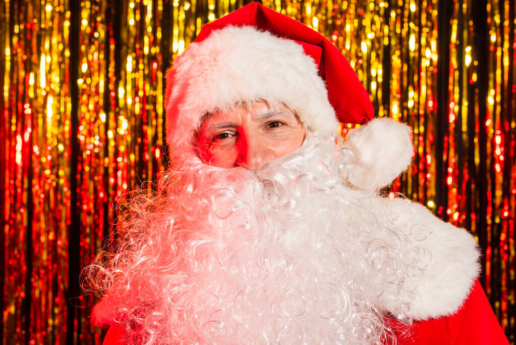 Портрет бородатого Санта-Клауса в шляпе, смотрящего в камеру рядом с размытой мишурой  - Фото, изображение