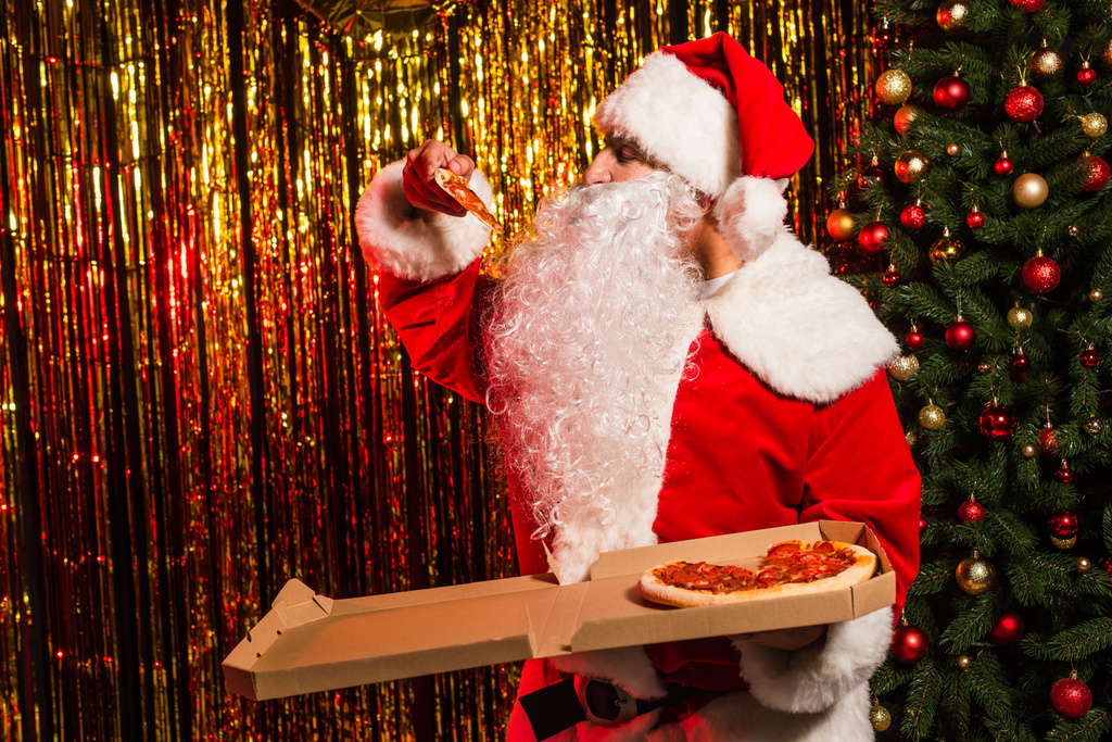 Санта-Клаус в костюме держит вкусную пиццу на вынос возле рождественской елки и мишуры  - Фото, изображение