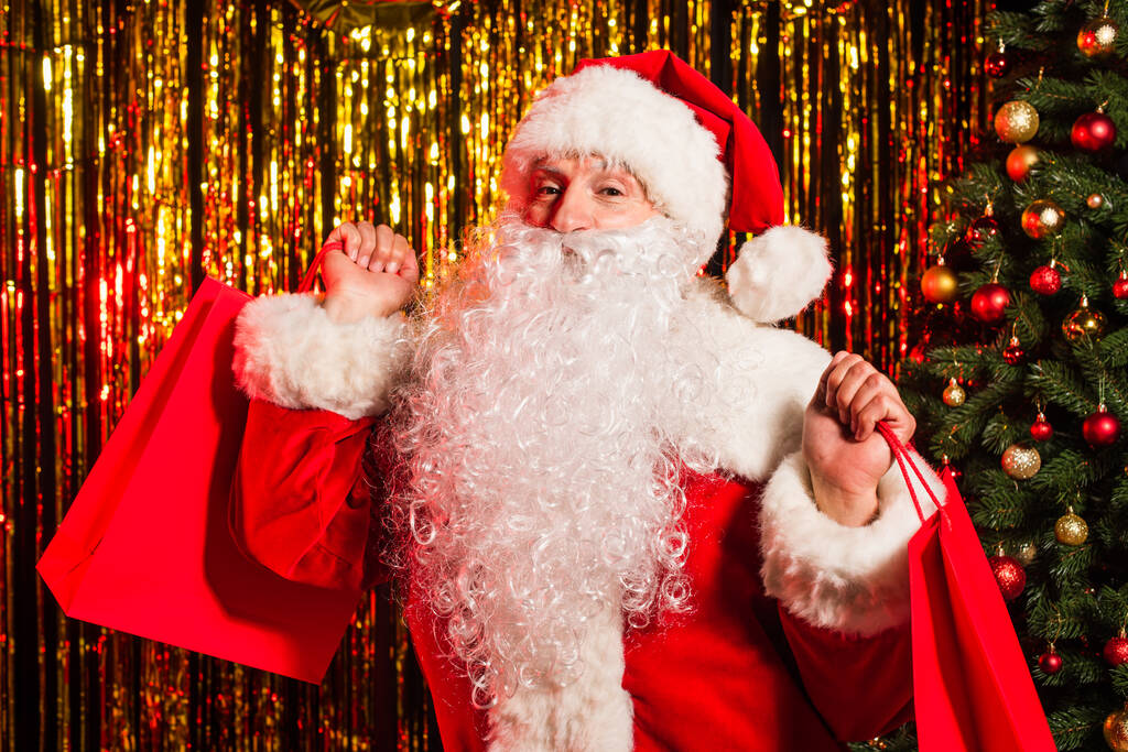 Санта-Клаус в костюме держит сумки для покупок возле рождественской елки и мишуры  - Фото, изображение