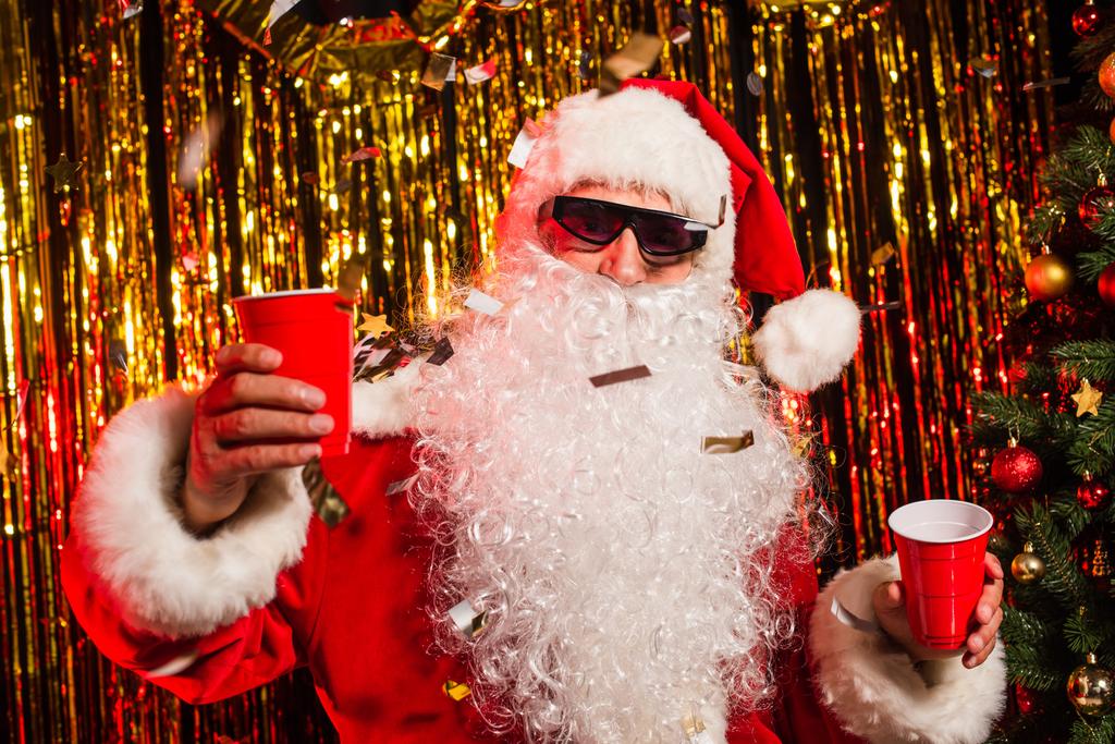 Weihnachtsmann mit Sonnenbrille hält Plastikbecher unter Konfetti in der Nähe des Weihnachtsbaums - Foto, Bild