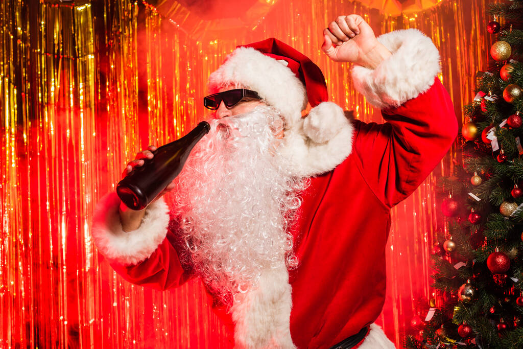 Père Noël dans des lunettes de soleil tenant bouteille de champagne près de l'arbre de Noël et tinsel  - Photo, image