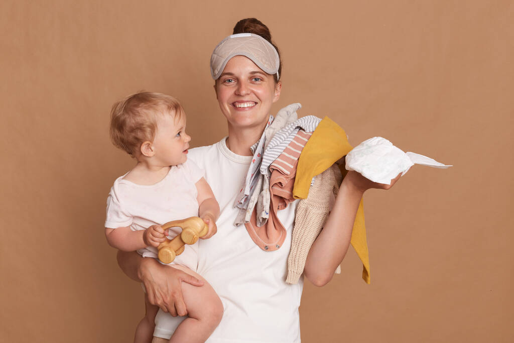 Χαμογελαστή νεαρή μητέρα με bun χτένισμα κρατώντας το παιδί της κόρη, τα ρούχα και την πάνα, κοιτάζοντας κάμερα με toothy χαμόγελο, απολαμβάνοντας άδεια μητρότητας της, θέτοντας απομονωμένη πάνω από καφέ φόντο. - Φωτογραφία, εικόνα